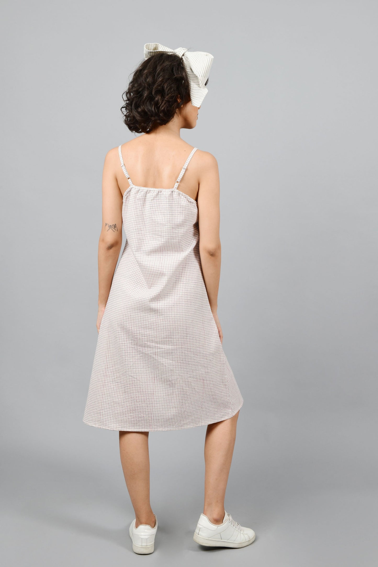 Model showing the back of the dress made by hand block printed handspun handwoven cotton khadi dress, printed with checks of red and blue on an off-white base. The model is also wearing an off-white printed bow in the same technique