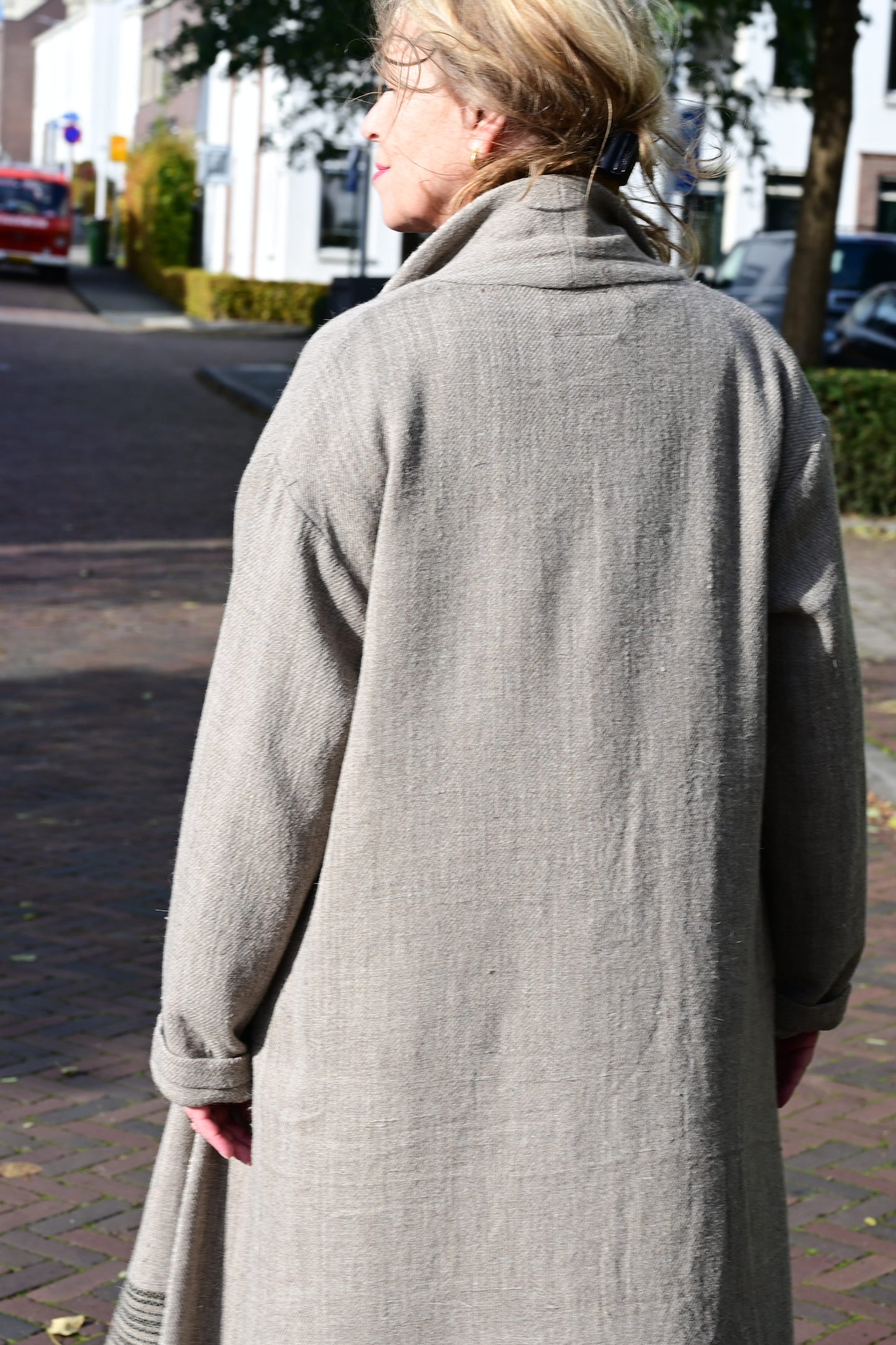 A middle aged european model posing with back towards the camera, wearing brown woolen long shrug with ivory woven pattern on the collar. This shrug is made of handspun and handwoven Indian sheep wool by Cotton Rack.