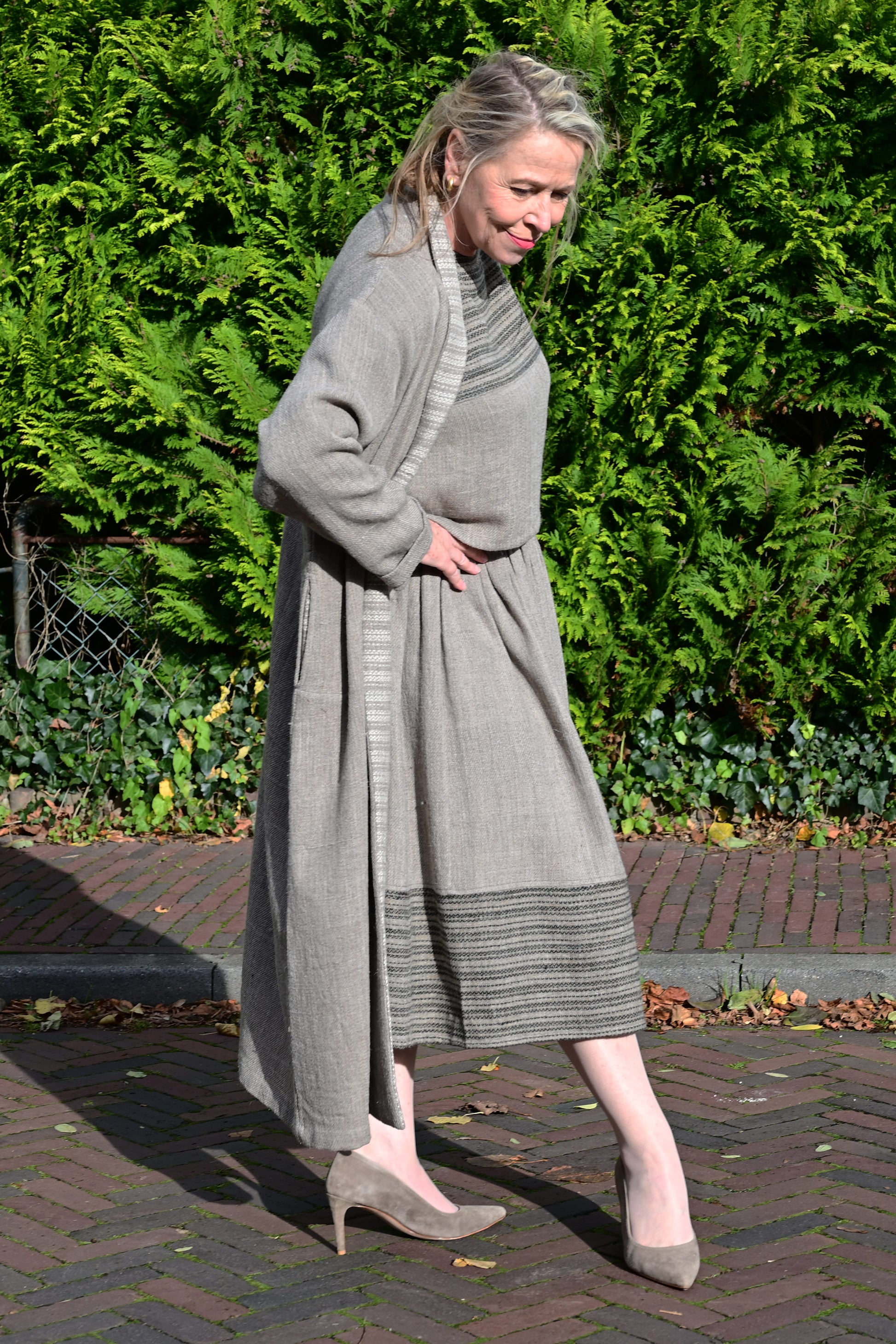 A middle aged european model posing wearing brown woolen long shrug with ivory woven pattern on the collar. This shrug is made of handspun and handwoven Indian sheep wool by Cotton Rack.