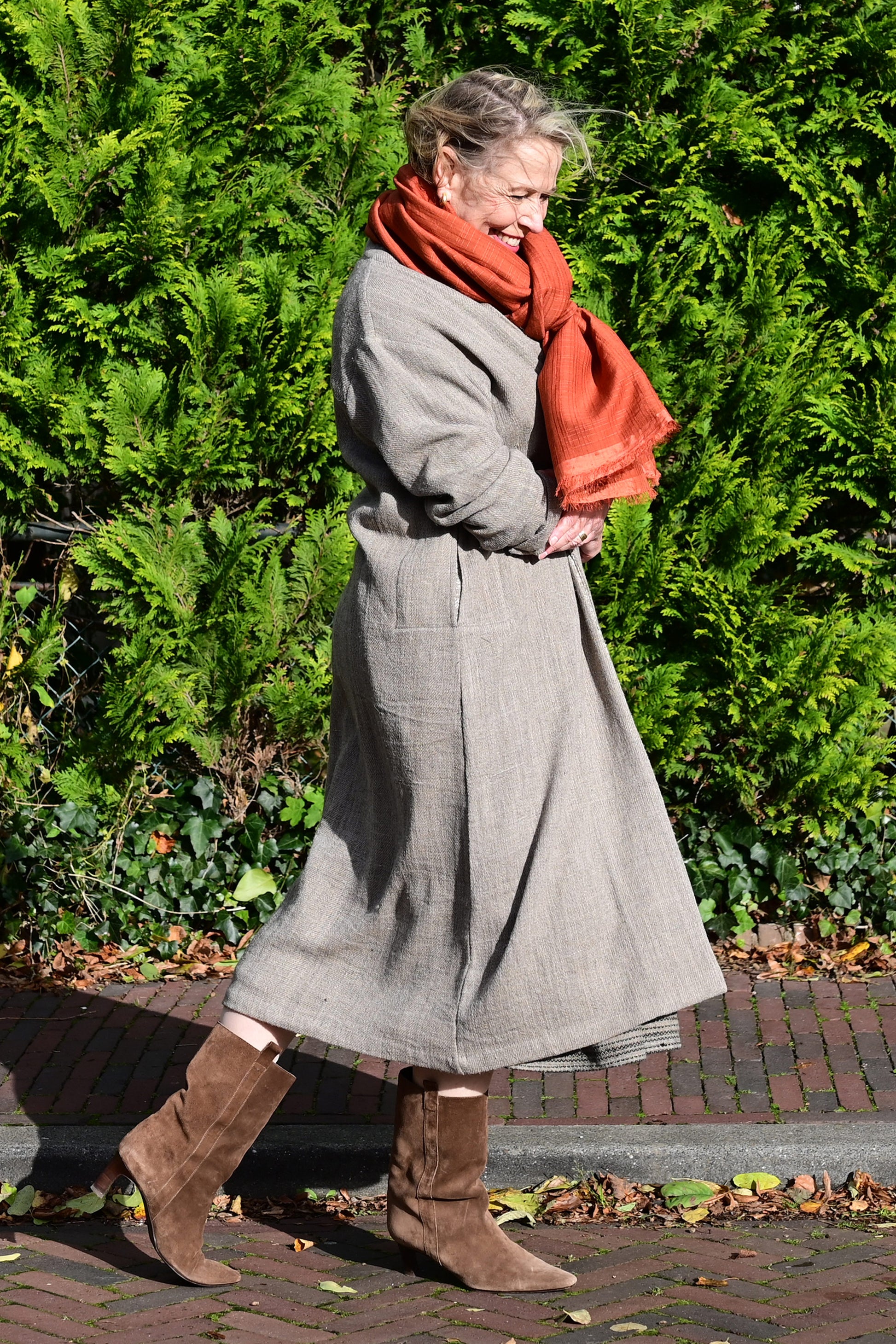 A middle aged european model walking on a street in netherlands wearing brown woolen long shrug with ivory woven pattern on the collar over a black wrap around dress. This shrug is made of handspun and handwoven Indian sheep wool by Cotton Rack.