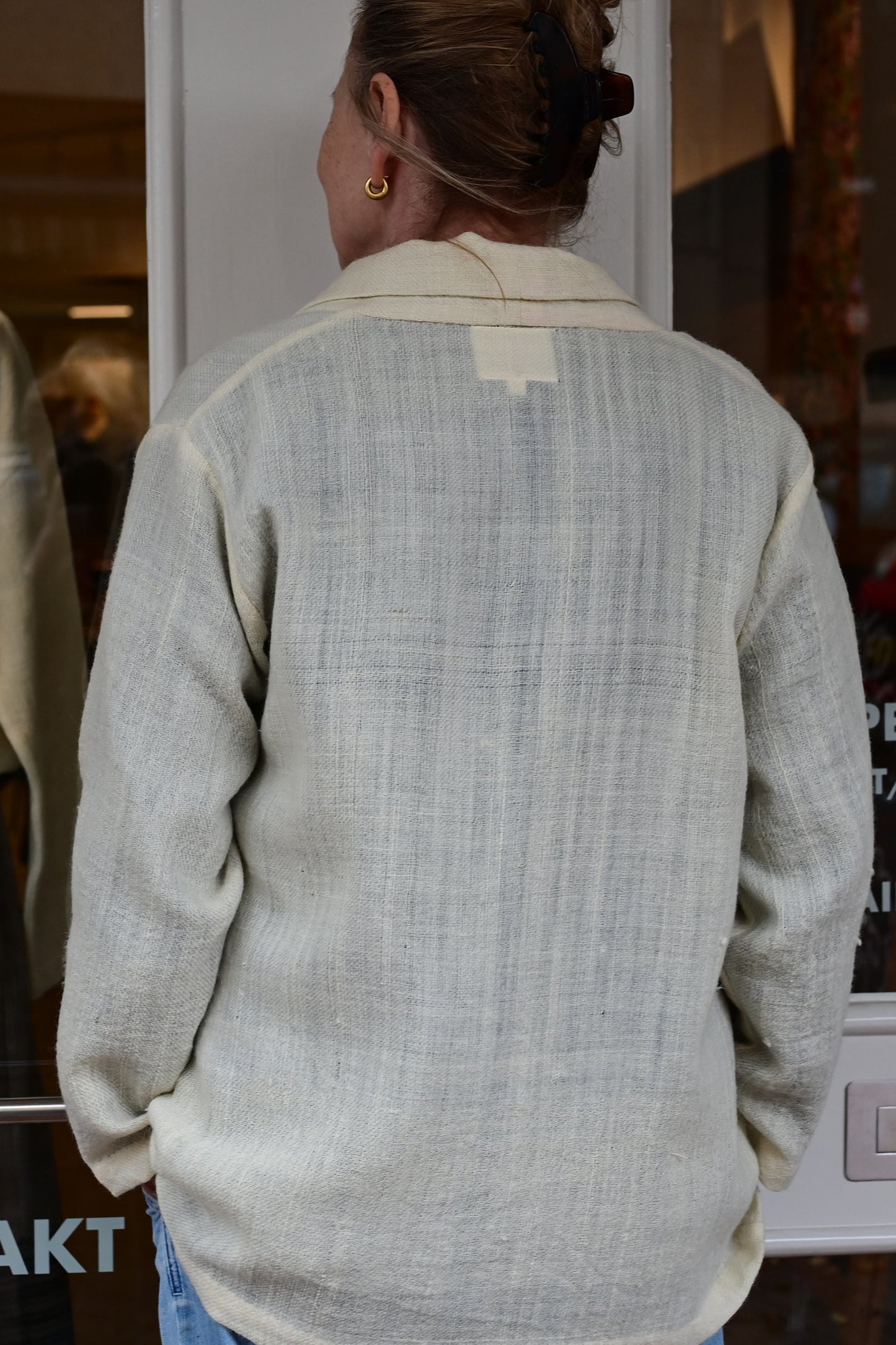 Back of a middle aged european model wearing an ivory white woolen cardigan shrug with long sleeves over blue washed denims. Shrug is made of handspun and handwoven Indian wool by Cotton Rack.