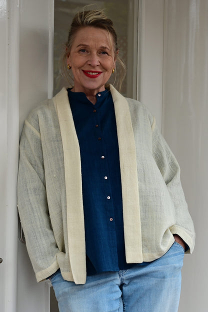 A middle aged european model wearing an ivory white woolen cardigan shrug with long sleeves over a blue silk shirt and blue washed denims. Shrug is made of handspun and handwoven Indian wool by Cotton Rack.