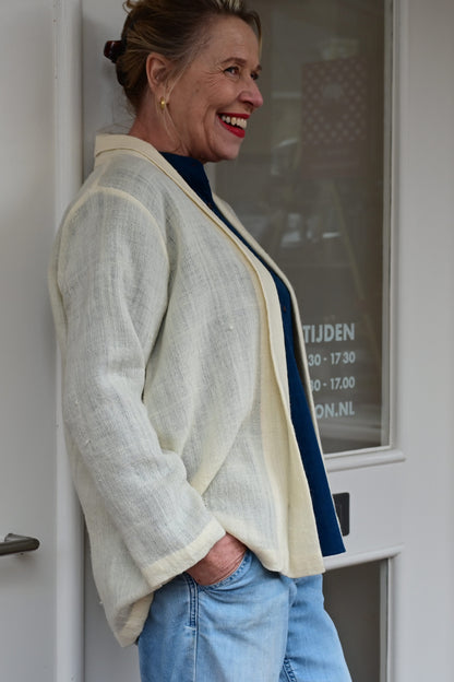 Side pose of a middle aged european model wearing an ivory white woolen cardigan shrug with long sleeves over a blue silk shirt and blue washed denims. Shrug is made of handspun and handwoven Indian wool by Cotton Rack.