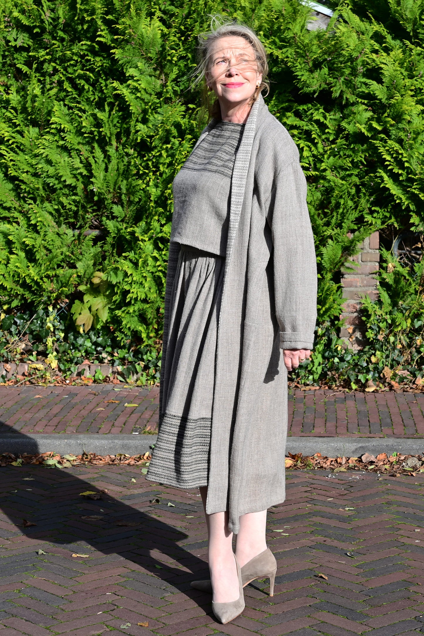 A middle aged european model posing wearing brown woolen long shrug with ivory woven pattern on the collar. This shrug is made of handspun and handwoven Indian sheep wool by Cotton Rack.