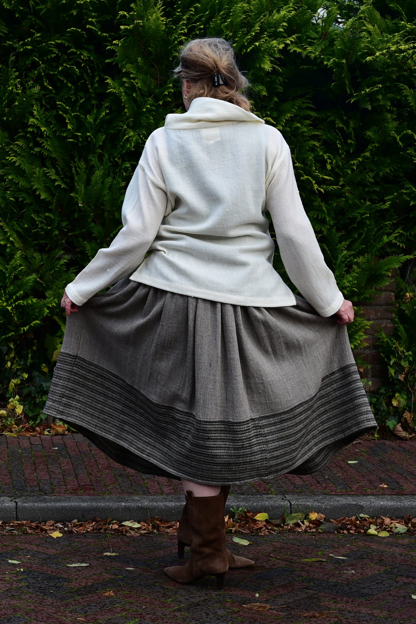 Standing back pose of a middle aged female model on a european street wearing an ivory white woolen sweater top and gathered skirt made of hand spun and handloom woven natural fabrics by Cotton Rack.